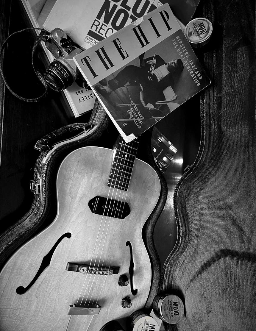 An open vintage guitar case with a blonde Gibson ES125, a film camera, Blue Note and The Hip books and MOJO Hair jars